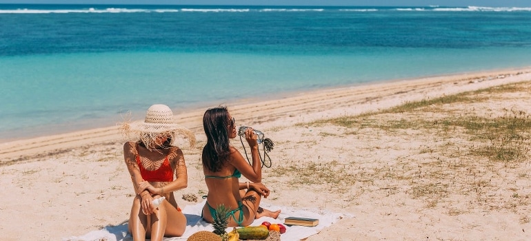 Two women in bikinis are sitting on the beach. 