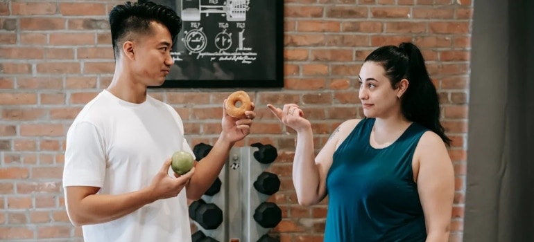 A personal trainer showing a woman two options a doughnut and an apple, a woman thinking about why nutrition matters as much as training