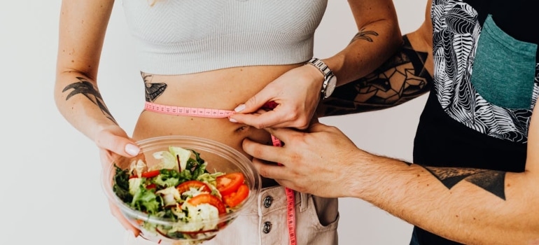 A man measuring a woman's waistline, a woman holding a bowl of salad