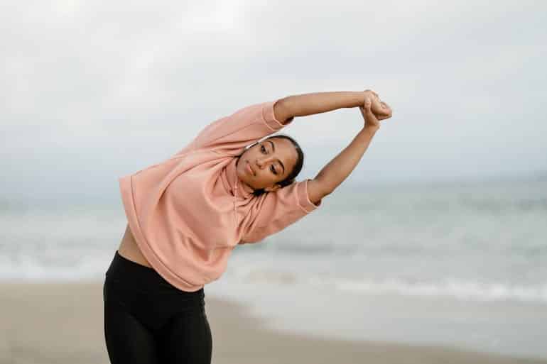 A woman in a pink sweater stretches on the beach, extending her arms as she performs a gentle warm-down near the shoreline.