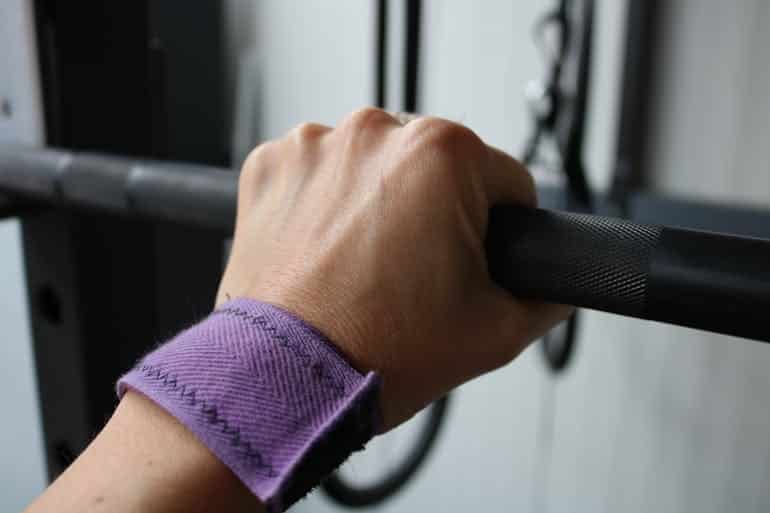 A close-up of a hand gripping a gym bar with a purple wrist wrap and blurred equipment in the background, showing why building grip strength improves more than just your lifts.