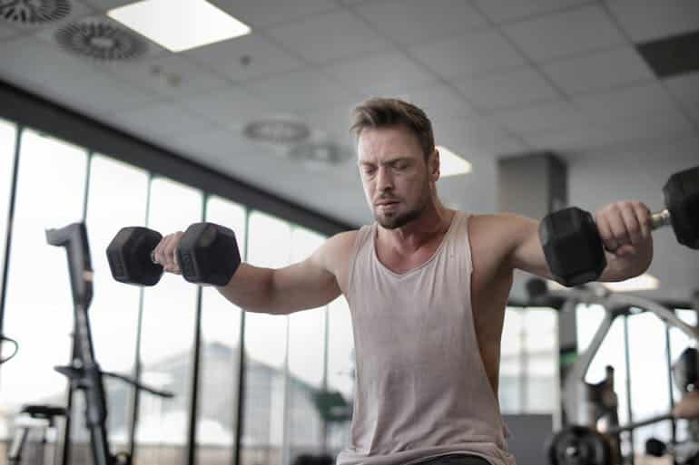 A person in a sleeveless shirt performs a weightlifting exercise with dumbbells in a bright gym setting with large windows.